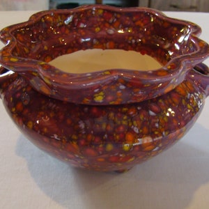 May include: A decorative ceramic bowl with a speckled, glossy finish. The bowl is primarily burgundy with orange, yellow, and white speckles. The rim has a scalloped edge. The interior is a light cream color.