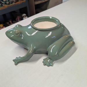 May include: A sage green ceramic frog-shaped planter with a smooth, glossy finish. The frog is depicted in a seated position with its front legs extended forward and back legs tucked. A circular opening with a cream-colored interior sits on its back.