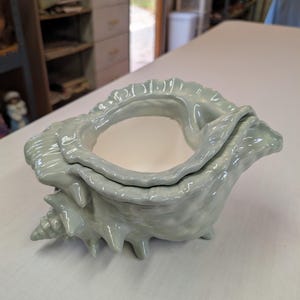 May include: A light blue ceramic seashell-shaped container with a white interior. The container has a textured surface, a spiral base, and a ruffled edge. The container is sitting on a white surface.