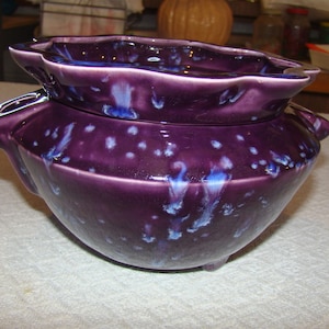 May include: A ceramic, purple-glazed decorative bowl with a lid. The bowl has a rounded shape with a ruffled rim and small handles. The glaze features white and blue speckles, adding visual interest. The bowl sits on three small feet.