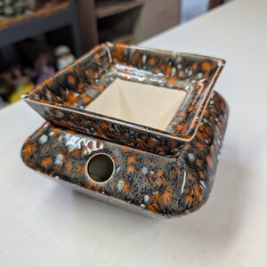 May include: A square ceramic wax warmer featuring a mottled glaze in shades of orange, black, and white. The top section is a removable dish with a cream-colored interior, while the base includes a circular opening on the front side for ventilation.