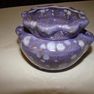 May include: Two-piece ceramic set in a deep purple hue, speckled with yellow and white accents. The top piece is smaller and sits atop a larger, bowl-shaped base. The edges have a wavy, scalloped design.