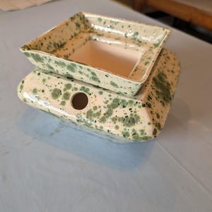 May include: A square ceramic wax warmer featuring a cream-colored base with a speckled green glaze. The top section is a removable square dish for wax melts, and the lower base includes a circular opening on the side for a power cord or light.