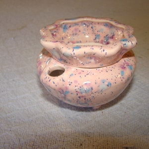 May include: A ceramic incense burner in a soft pink hue, speckled with blue and purple accents. The burner features a scalloped top and a rounded base with a small hole, perfect for holding incense sticks. The overall design is whimsical and decorative.