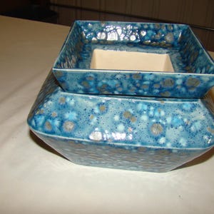 May include: A two-piece ceramic planter set. The top piece is a square tray, and the bottom piece is a larger, geometric-shaped base. Both pieces are glazed in a speckled blue and gray pattern, creating a textured appearance. The interior of the top piece is a cream color.