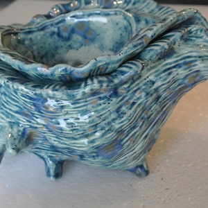 May include: A ceramic seashell-shaped bowl with a glossy finish. The bowl is primarily turquoise with blue and gold accents. It has a textured surface and sits on three small feet. The design mimics the natural form of a seashell.