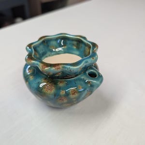 May include: A small, round ceramic oil burner featuring a glossy teal glaze with brown crystalline speckles. The top section has a scalloped, wavy rim, while the base includes a small circular opening on the side for airflow or access.