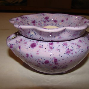 May include: A two-piece ceramic container set in a light purple hue, speckled with dark purple and blue dots. The top piece has a scalloped edge, and the bottom has small handles. The set is designed for decorative use.