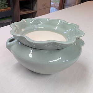 May include: A two-piece ceramic pottery set in a soft, muted green. The bottom piece is a rounded bowl with small handles, and the top is a scalloped-edge dish. The interior of both pieces is a creamy white color. The set is placed on a white surface.