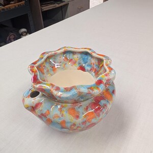 May include: A ceramic bowl with a scalloped rim and a rounded base, featuring a vibrant multicolored glaze of blue, orange, red, and yellow speckles. The interior of the bowl is a smooth, solid cream color, and a small circular opening is visible.