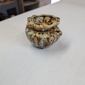May include: A small, two-piece ceramic oil warmer featuring a scalloped top bowl and a rounded base. The piece is finished with a marbled glaze in shades of brown, tan, and light blue, creating a swirling, organic pattern across the entire surface.