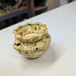 May include: A ceramic oil warmer with a pale yellow glaze featuring dark brown and blue speckles. The piece consists of a rounded base with a small circular opening and a removable top dish with a scalloped, flower-like rim edge.