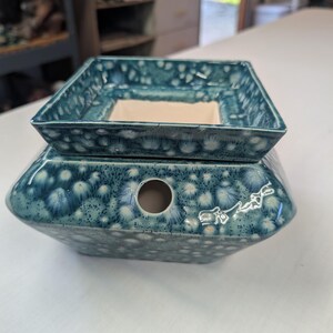 May include: A square ceramic wax warmer featuring a teal and blue crystalline glaze with white starburst patterns. The base has a circular opening on the side and a signature etched into the glaze. A removable square dish sits on top.