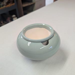 May include: A round, sage green ceramic bowl with a smooth, glossy finish. The bowl features a wide, open top with a thick, rounded rim and a small, semi-circular notch cut into the side of the rim, revealing a light cream-colored interior.