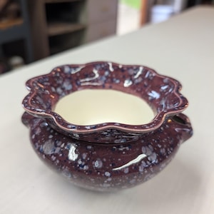 May include: A ceramic bowl with a scalloped rim and two small side handles. The exterior features a dark plum glaze with a speckled pattern of white and light blue dots, while the interior is a smooth, solid off-white color.