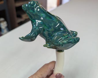 May include: A ceramic garden stake featuring a glossy, dark green and teal mottled frog figurine perched atop a smooth, cylindrical white stem. The frog is sculpted in a seated position with its head tilted upward, showcasing a reflective glaze.