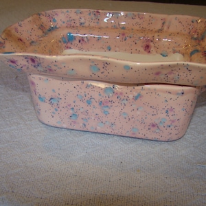 May include: Two-piece ceramic serving dish set. The set features a light pink base color with a speckled pattern of blue and purple dots. The top dish has a ruffled edge.
