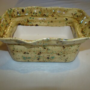 May include: A rectangular ceramic planter with a speckled beige glaze, accented with brown and teal spots. The rim has a wavy, scalloped edge. The interior is white, creating a contrast with the exterior.