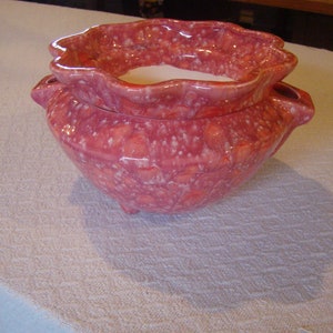 May include: A ceramic planter with a ruffled rim and a speckled, reddish-pink glaze. The planter has a rounded body, three small feet, and two side handles. The interior is white.