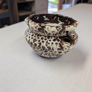 May include: A two-piece ceramic oil warmer featuring a speckled cream and dark brown glaze. The top section is a shallow bowl, while the bottom base is a rounded vessel with a small circular opening on the side for a heat source.