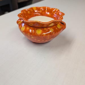 May include: A ceramic wax warmer featuring a vibrant orange and yellow mottled glaze. The bowl has a scalloped, ruffled edge and a smooth, cream-colored interior basin. The base is rounded with small side handles and a small opening on the side.