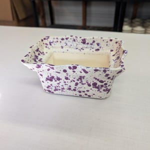 May include: A square ceramic planter with a cream-colored interior and exterior. The exterior is decorated with purple speckles. The planter has a wavy rim and is sitting on a white surface. The background shows shelves with other ceramic items.