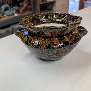May include: A two-piece ceramic planter set. The black base is speckled with red, orange, and green spots. The top piece has a scalloped edge and a white interior. The planter sits on a white surface.
