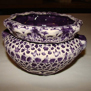 May include: Two ceramic bowls, one stacked on the other. The bowls are a deep purple color with a textured, lace-like pattern in white. The rims have a scalloped edge. The bowls are likely for decorative or functional use.