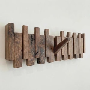 May include: A wall-mounted coat rack constructed from dark brown wood with a natural grain pattern. The design features multiple vertical rectangular slats of varying lengths, with one angled wooden peg extending outward for hanging items.