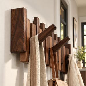 Puede incluir: Un perchero de pared fabricado en madera de color marrón oscuro, con múltiples listones verticales de alturas variables y varios ganchos inclinados hacia afuera. Una bufanda de tela beige y un bolso de cuero tostado cuelgan de los ganchos.