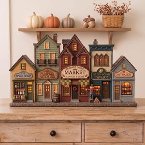 Könnte beinhalten: Ein dekoratives Tischdorf-Display mit fünf verbundenen Gebäuden: The Spice Shop, Pumpkin Patch Inn, Orchard Grove Market, Gilded Acorn Tavern und Cozy Bakery. Die Strukturen sind in gedeckten Erdtönen gehalten und haben beleuchtete Fenster.