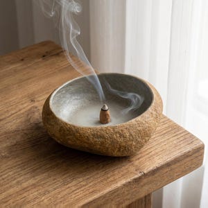 Peut inclure: Un brûleur d'encens en forme de pierre ronde avec un extérieur beige texturé et rugueux et un bassin intérieur gris clair lisse. Un petit cône d'encens marron est placé au centre, émettant un fin filet de fumée blanche qui s'élève en tourbillons.