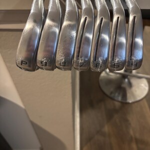 May include: A set of silver golf clubs, including a pitching wedge and irons numbered 9, 8, 7, 6, and 5. The clubs are arranged in a row, with the club heads facing the viewer. The clubs are new and still wrapped in plastic.