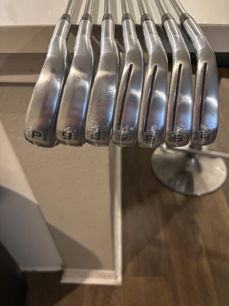 May include: A set of silver golf clubs, including a pitching wedge and irons numbered 9, 8, 7, 6, and 5. The clubs are arranged in a row, with the club heads facing the viewer. The clubs are new and still wrapped in plastic.