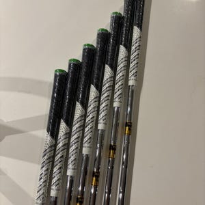 May include: A set of golf clubs with black grips and white and black patterned shafts. The grips have green accents. The clubs are arranged in a row, with the club heads out of frame. The clubs are on a white surface.