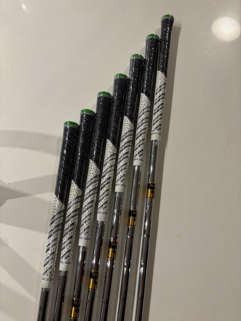 May include: A set of golf clubs with black grips and white and black patterned shafts. The grips have green accents. The clubs are arranged in a row, with the club heads out of frame. The clubs are on a white surface.
