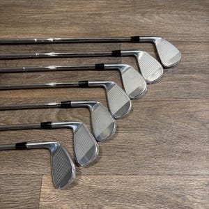 May include: A set of six shiny, silver golf irons arranged diagonally on a wooden surface. The clubs have black grips and grooved faces. The golf clubs are designed for precision and distance.