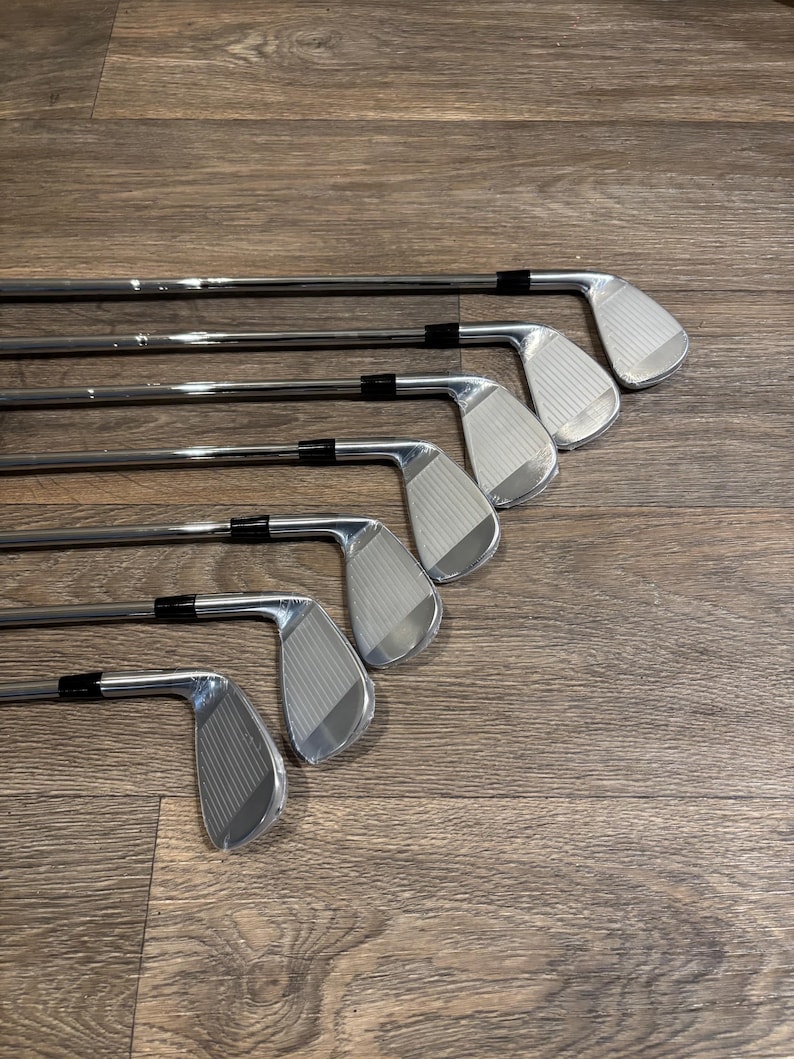 May include: A set of six shiny, silver golf irons arranged diagonally on a wooden surface. The clubs have black grips and grooved faces. The golf clubs are designed for precision and distance.