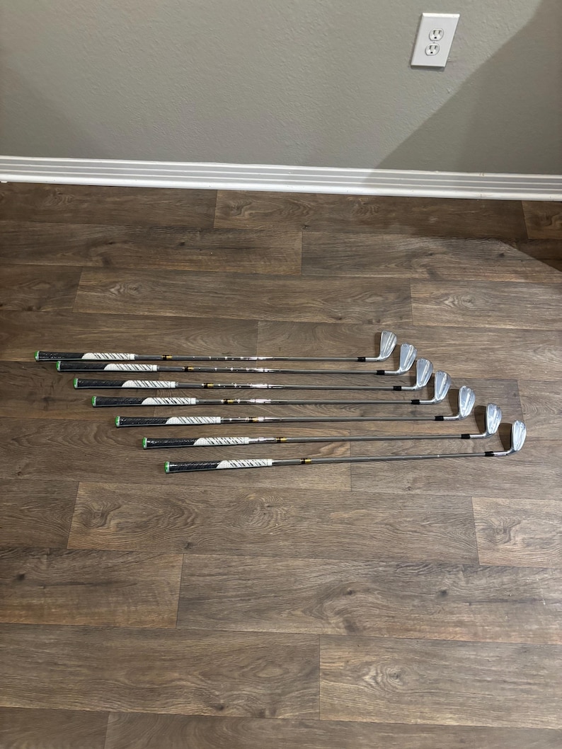 May include: A set of golf clubs arranged on a wooden floor. The clubs have black grips with white and silver accents, and silver club heads. The clubs are angled to the right, creating a visual flow.