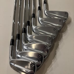 May include: A set of shiny, silver golf irons arranged in a row. The clubs have black grips and the brand name "TaylorMade" is visible. The club heads have the model name "D62.i" etched on them.