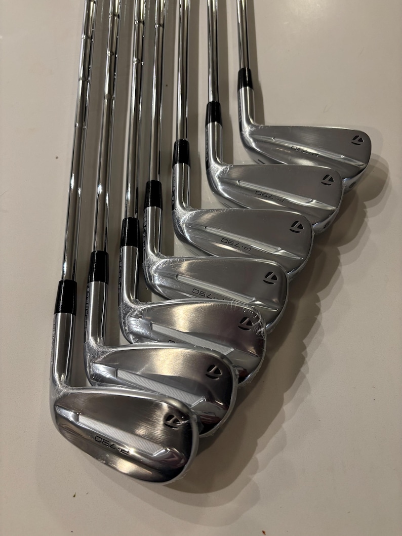 May include: A set of shiny, silver golf irons arranged in a row. The clubs have black grips and the brand name "TaylorMade" is visible. The club heads have the model name "D62.i" etched on them.