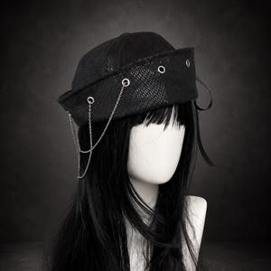 Può includere: Un cappello in stile marinaio nero con un motivo testurizzato simile alla pelle di serpente sulla tesa. Il cappello è decorato con occhielli in metallo argentato e una sottile catenella argentata drappeggiata sul lato. Un nastro nero pende dietro.