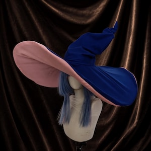 Può includere: Un grande cappello da strega a tesa larga con un esterno blu intenso e una parte inferiore a contrasto color rosa pallido. Il cappello è esposto su una testa di manichino con capelli blu lisci alle spalle, che mette in risalto la corona.