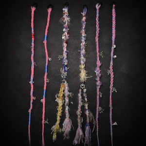 May include: Six decorative hair wraps featuring various textures, colours, and charms. The wraps include braided pink, blue, and multi-coloured yarn, frayed fabric strips, and metallic accents like small silver-toned charms and beads attached throughout.