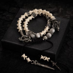 May include: A bracelet featuring a series of off-white bone-like vertebrae segments, grey stone beads, and intricate silver-toned metal dragon-shaped charms. The piece includes a black cord with adjustable ends and a silver-toned metal spike-shaped pendant.