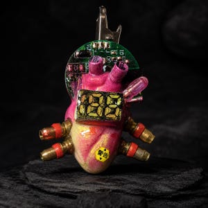 May include: A sculpted anatomical heart ornament featuring a green circuit board top, a central digital display showing 88, and four brass-toned metal ports with red accents. A small yellow triangular warning label with black Japanese text is visible.