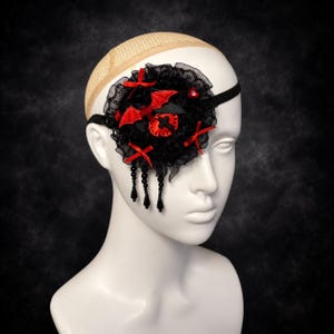 May include: A black lace eye patch featuring red satin bows, a red bat emblem, and a central red heart with a black cross. The accessory is adorned with dangling black beads and is secured with a black elastic band around a white mannequin head.