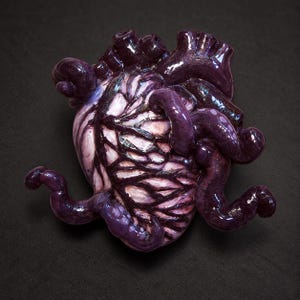 May include: A glossy, sculptural anatomical heart figurine featuring a pale pink base with deep purple, raised vein-like structures and tubular extensions. The surface has a reflective, polished finish with dark purple accents throughout the form.