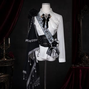 Peut inclure: Une chemise blanche à manches longues avec un corset noir à la taille, une écharpe décorative à motif floral, une rose noire et un drapé en tissu noir orné de dentelle argentée et de perles en forme de cristaux suspendues.