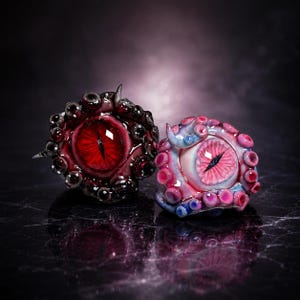 May include: Two sculpted eye-shaped ornaments featuring intricate tentacles and glossy finishes. One piece is deep black with a vibrant red iris and black pupil, while the other is light pink and blue with a matching pink iris and black pupil.
